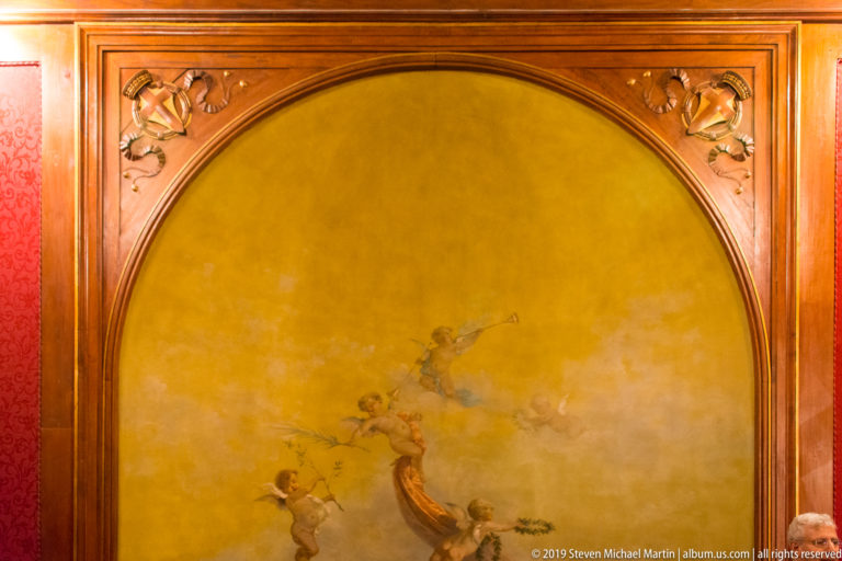 Arched golden wall painting with small winged figures, enclosed by polished wood molding with carved floral details.