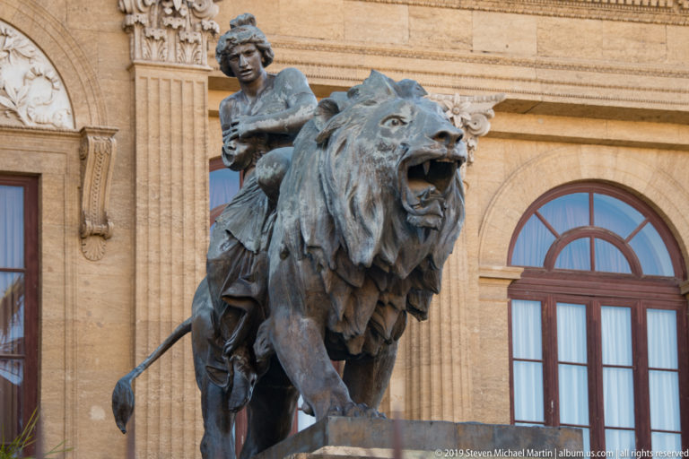 Bronze lion with open mouth and seated female figure in front of the Teatro Massimo façade.