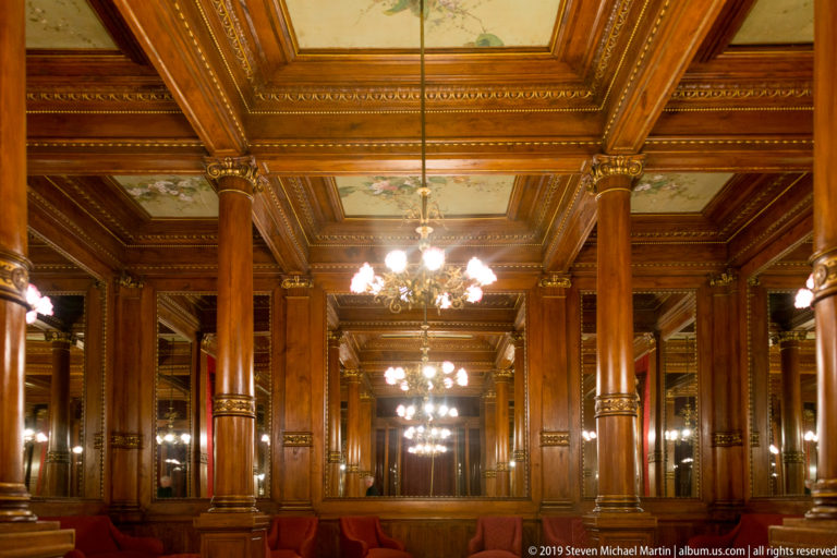 Long wood-paneled salon with repeated columns, mirrors, chandeliers, and pale painted ceiling panels receding into the distance.