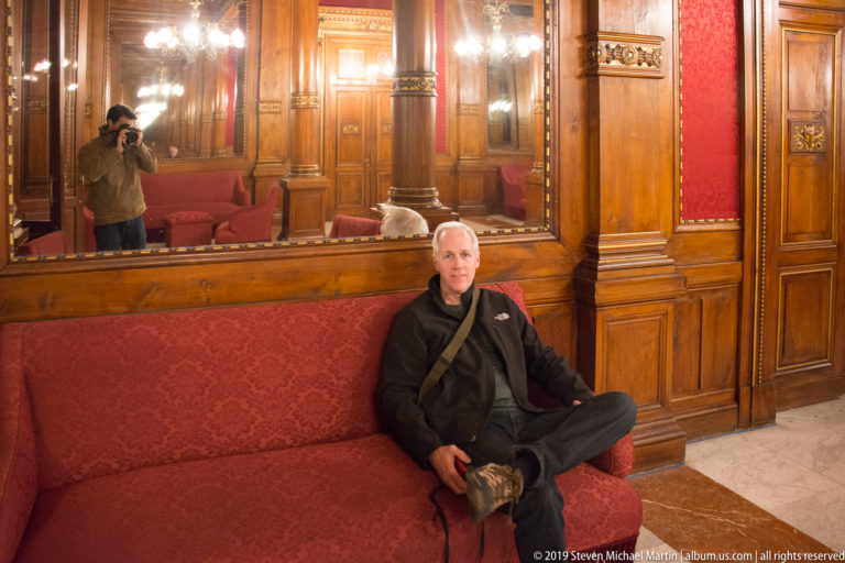 Visitor seated on a long red sofa in a polished wood-paneled theater salon with mirrors and carved details.