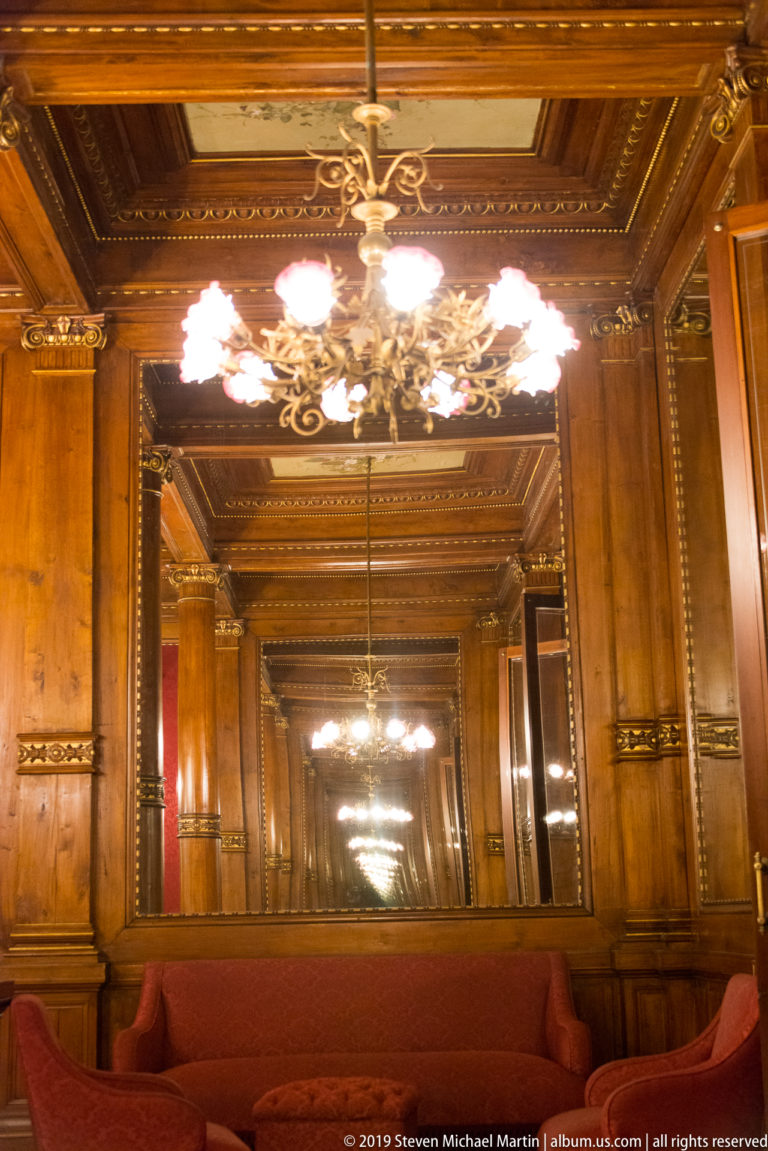 Large mirror reflects chandeliers, polished wood paneling, and red upholstered seating in an ornate theater salon.