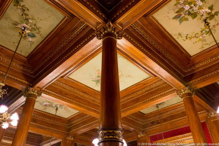 Polished wooden columns support a coffered ceiling with pale floral-painted panels, dark beams, and gilded decorative accents.