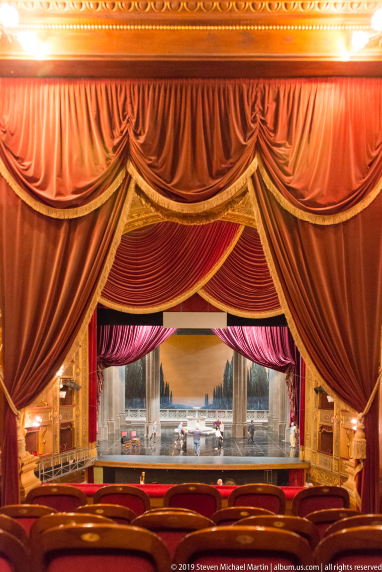 Rows of box seats face an open stage framed by layered red and burgundy curtains viewed from a private box.