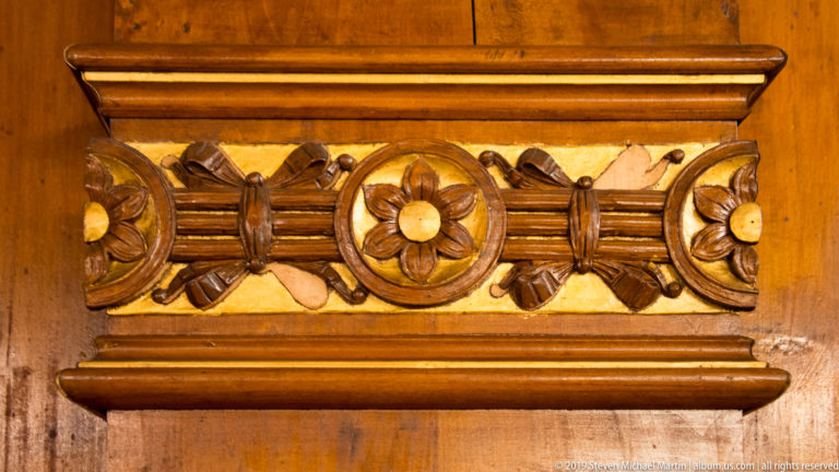 Close-up of polished wood paneling carved with circular flower rosettes, folded ribbon motifs, and pale decorative inlays.