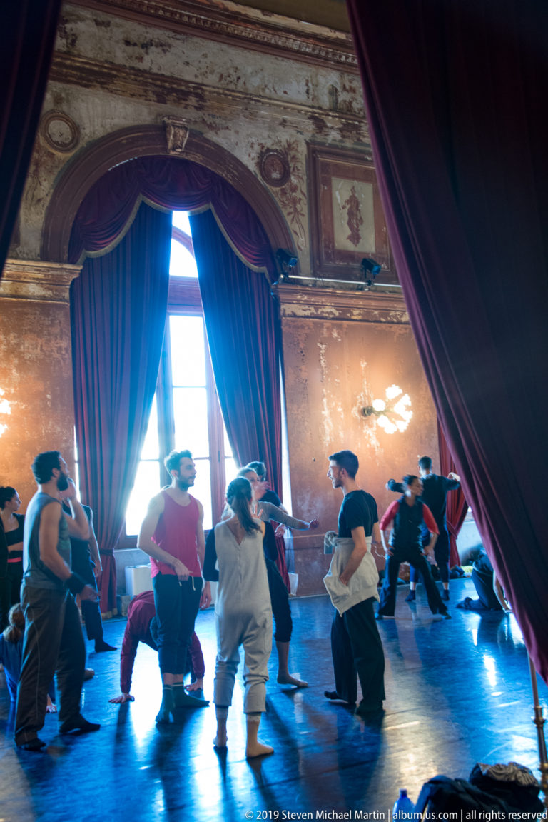 Backlit performers rehearse beside a tall window framed by crimson curtains in an ornate theater salon.