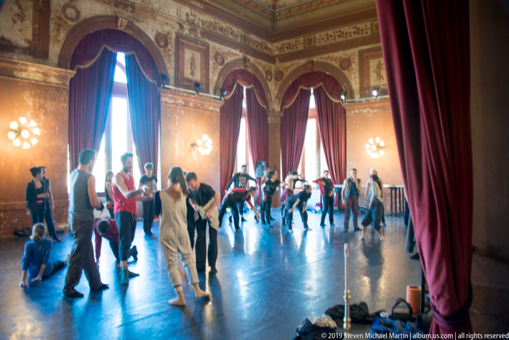 Performers rehearse on a polished floor in an ornate salon with tall red curtains, painted walls, and bright wall lights.