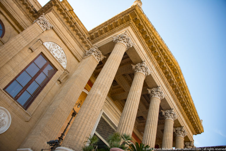 Low-angle view of tall fluted Corinthian columns, entablature, window, and sculptural roundel on the theater façade.