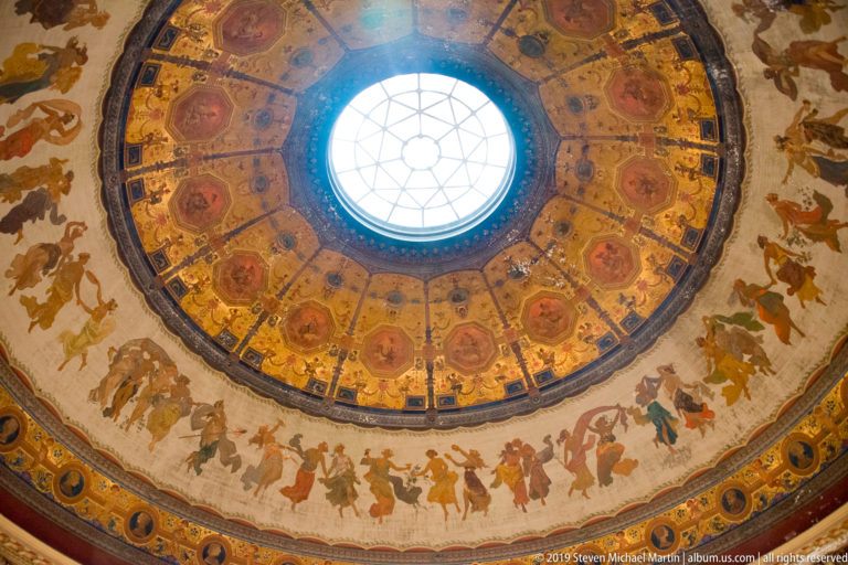 Central round skylight surrounded by radial painted ceiling compartments, gold ornament, and a broad figurative frieze.