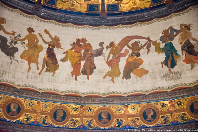 Painted procession of colorful figures around a curved frieze above blue-and-gold portrait medallions.
