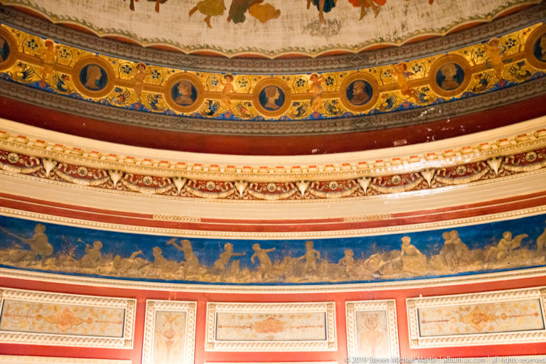Close view of blue, gold, red, and white decorative bands with profile medallions, classical molding, and gilded figures.