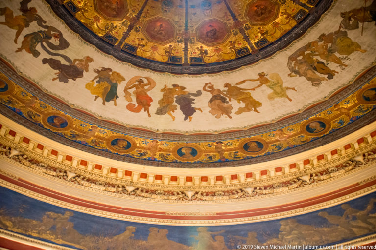 Curved interior frieze with painted figures above blue-and-gold portrait medallions and layered ornamental moldings.