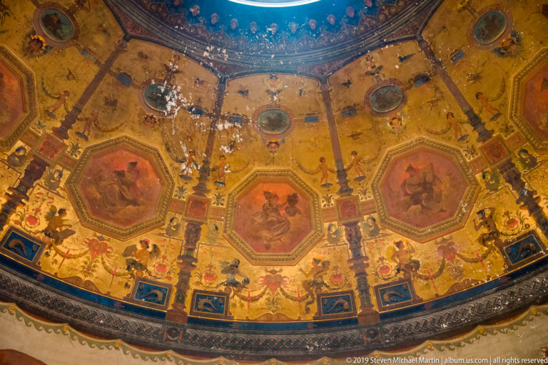 Ornate circular ceiling painted in gold tones with octagonal figurative panels, decorative borders, roundels, and visible surface wear.