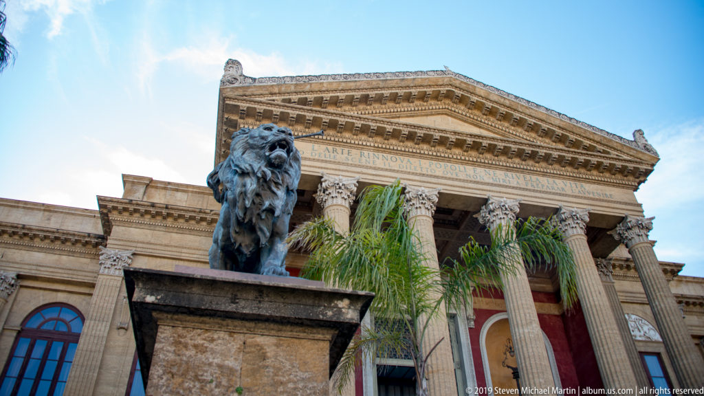 Bronze lion sculpture on a stone pedestal before the columned entrance and inscribed pediment of Teatro Massimo.