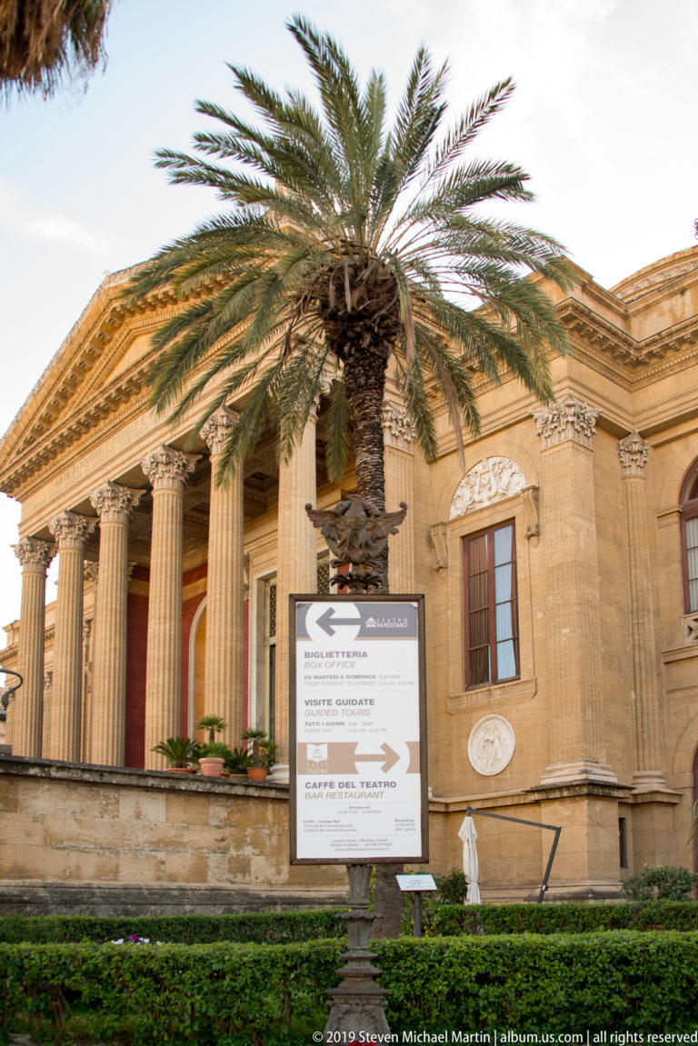 Palm tree and visitor sign in front of Teatro Massimo’s pale stone portico with tall classical columns and pediment.