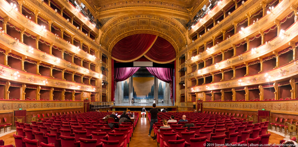Red auditorium seats face the open stage, surrounded by illuminated tiers of gilded theater boxes and draped curtains.