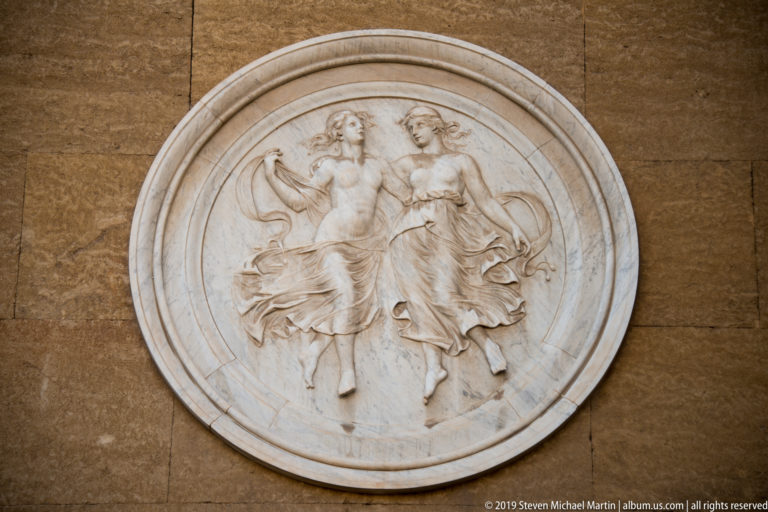 Round pale stone relief with two draped female figures mounted on a tan masonry wall.