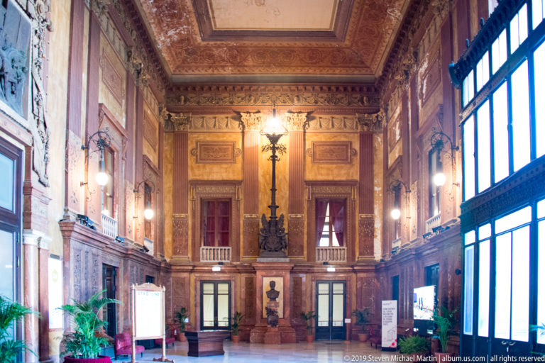 Grand theater hall with decorated ceiling, stone wall panels, lamps, portrait bust, and tall glazed windows.