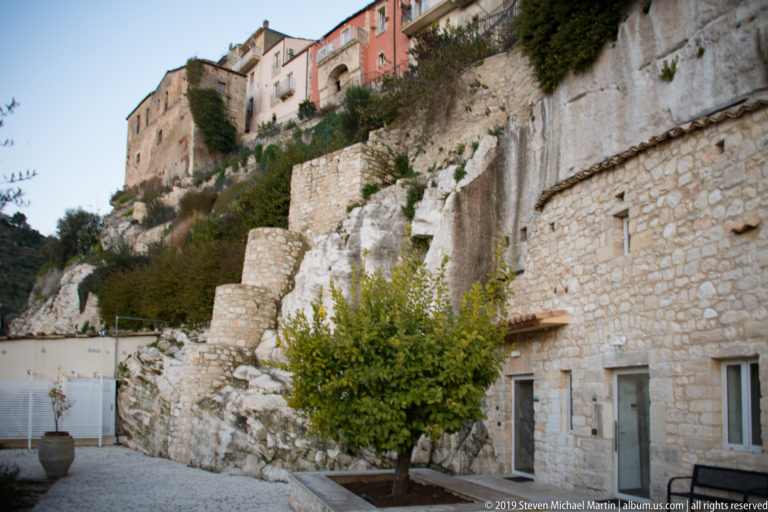 Historic Ragusa buildings perched above exposed pale limestone, terraced retaining walls, vegetation, and lower masonry structures.
