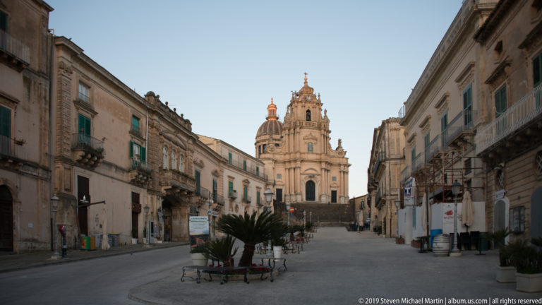 Historic Ragusa street with balconies and green shutters leading toward an elevated Baroque church at the far end.