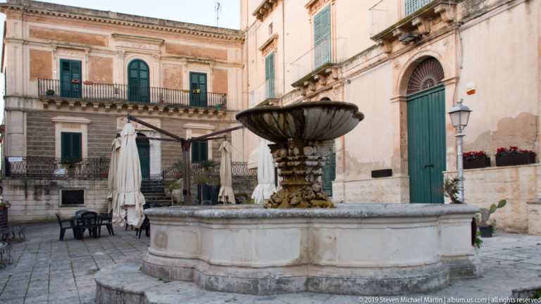 Large circular stone fountain with carved pedestal and dark scalloped bowl in a small square bordered by historic buildings.