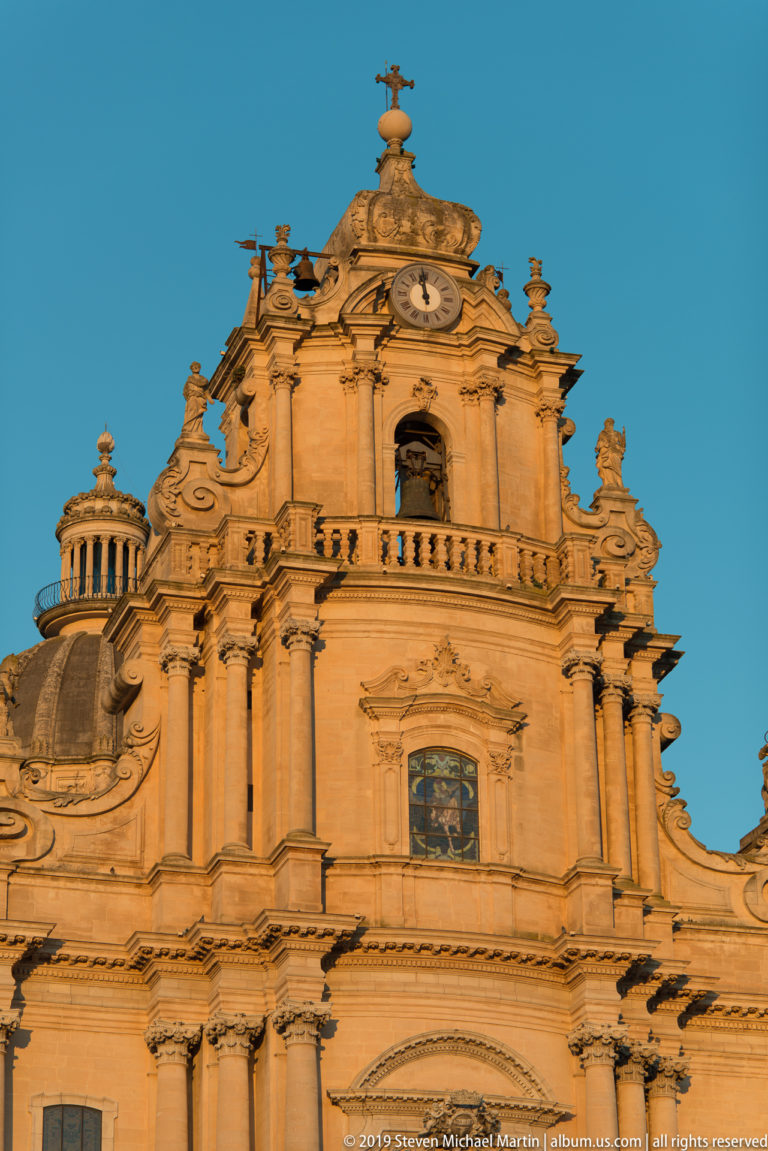 Monumental pale stone Baroque church façade with giant columns, clock, sculptural finials, upper balcony, and dome at left.