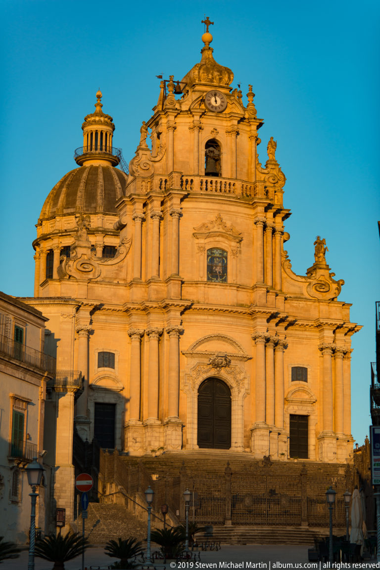 Full frontal view of a richly articulated pale stone Baroque church with central doorway, giant columns, clock, cross, and dome at left.