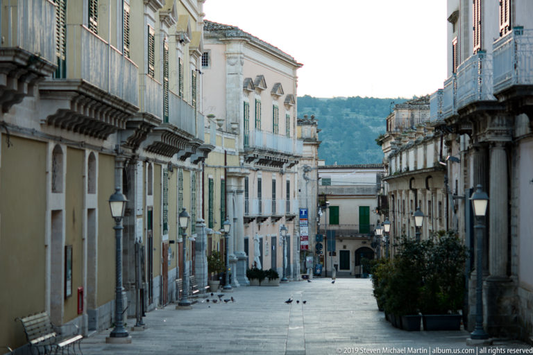Broad paved Ragusa street lined with historic palazzi and balconies, with pigeons, planters, lamps, and a wooded hillside beyond.