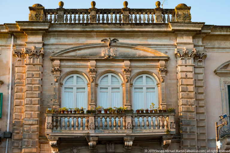 Ornate pale stone façade with three arched windows, carved pilasters, flowered balustraded balcony, rusticated corners, and rooftop balustrade.