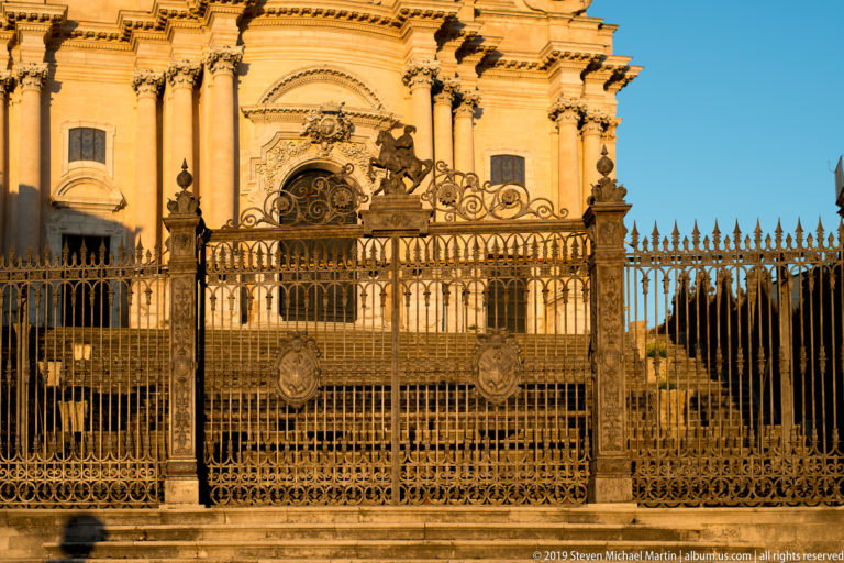 Ornate iron fence and gate with medallions in front of a broad stair and richly decorated pale stone church façade.