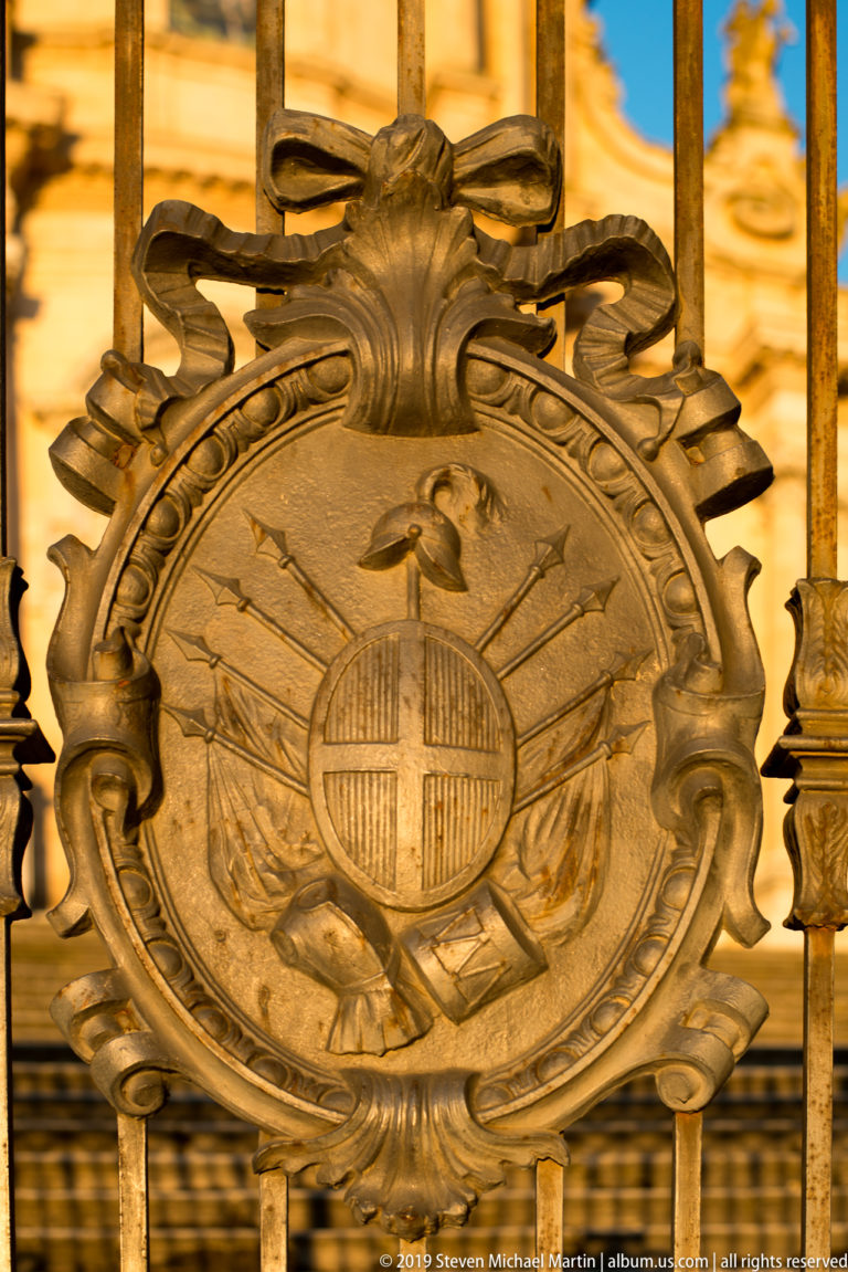 Close view of an oval heraldic-style medallion with crossed implements and scrolling ornament set into an iron gate.