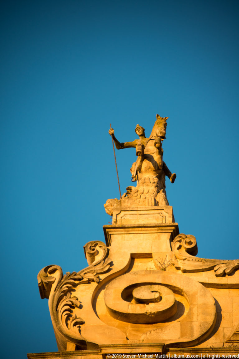 Stone equestrian saint on a rearing horse above a richly scrolled Baroque pediment against a clear blue sky.