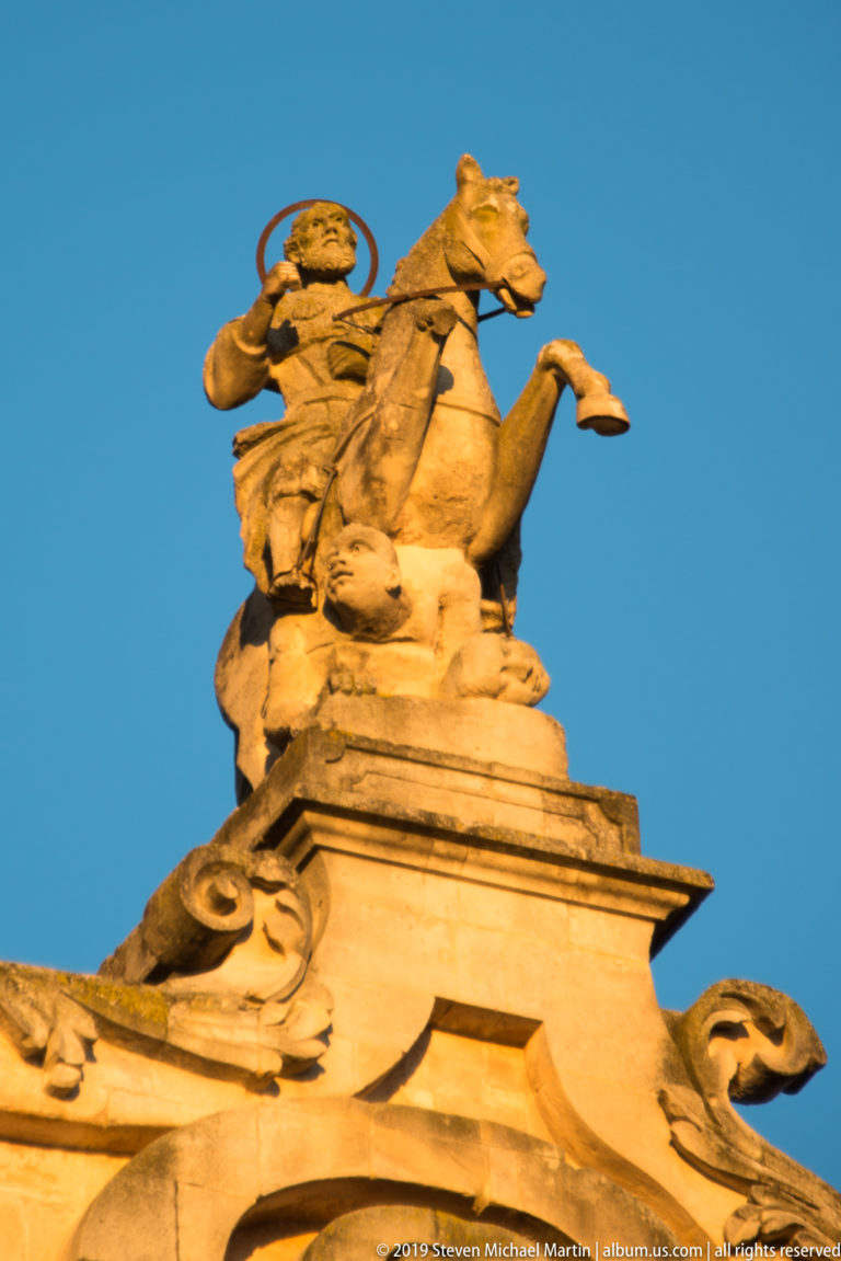 Stone sculpture of Saint George on a rearing horse above a dragon, mounted high on a church façade against blue sky.