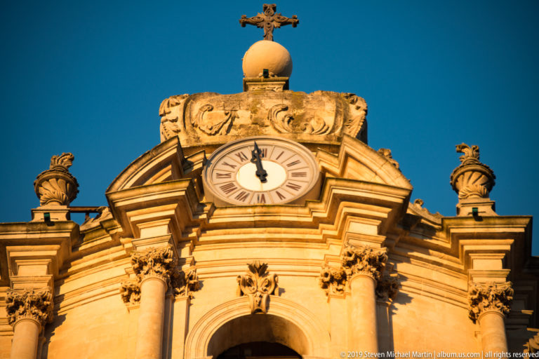 Ornate Baroque church pediment with round Roman-numeral clock, columns, urn finials, scrollwork, sphere, and cross.