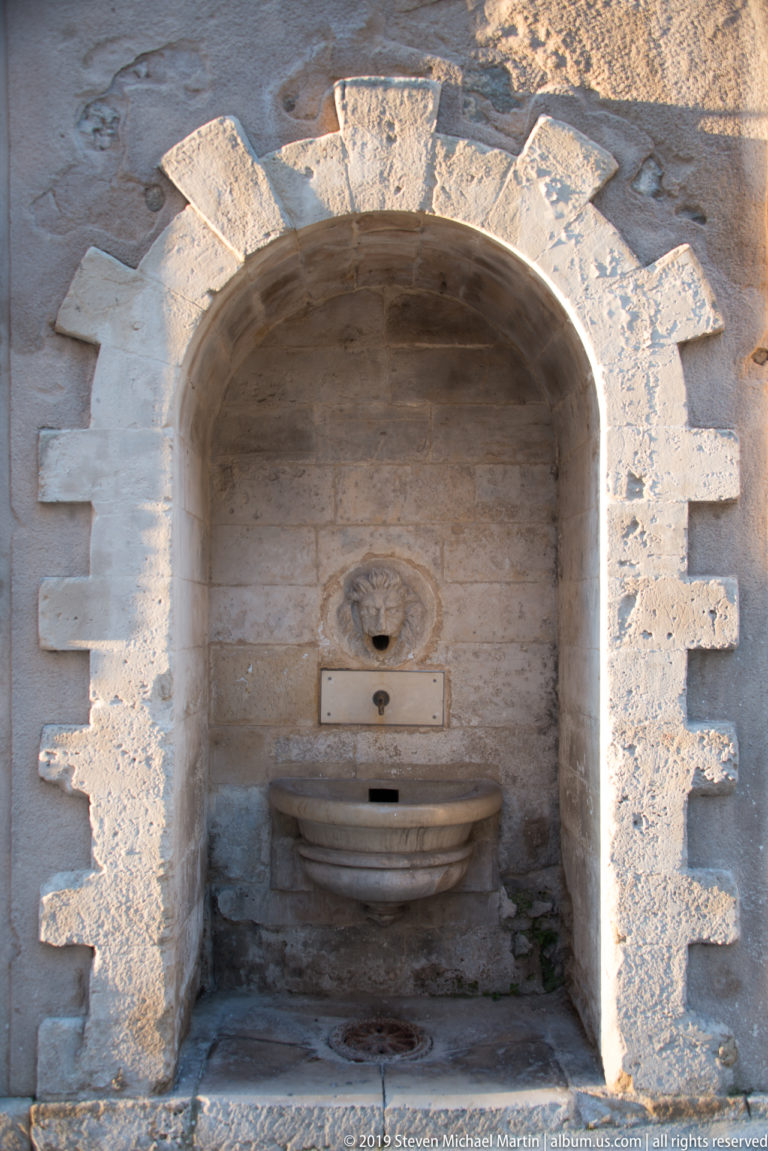 Small stone wall fountain with circular spout and projecting basin recessed within a deep arched masonry niche.