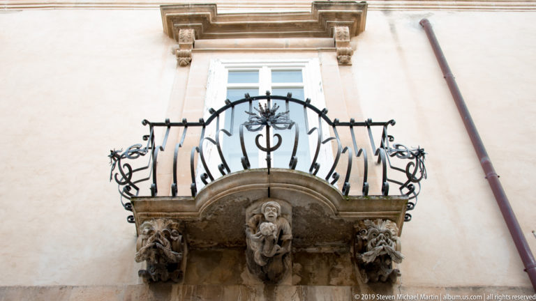 Upward view of a small wrought-iron balcony supported by a central carved human figure and flanking sculptural stone brackets.