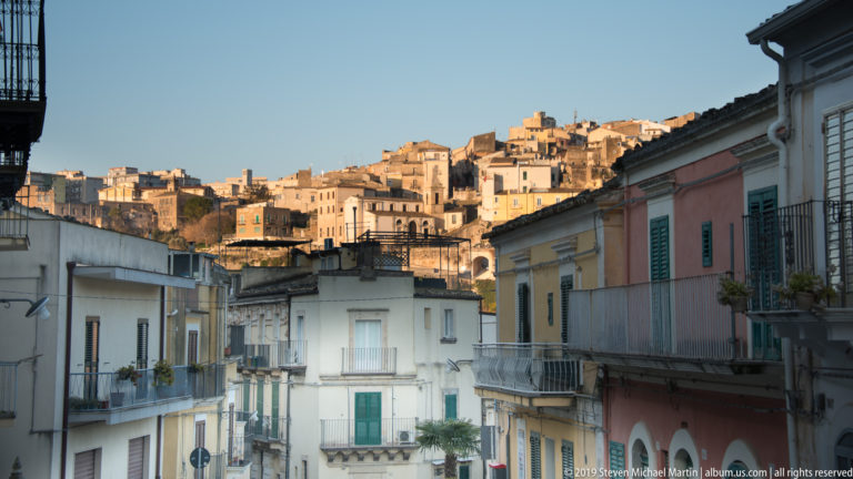 Pastel residential buildings with balconies and green shutters in the foreground, with Ragusa’s sunlit hillside quarter rising behind.