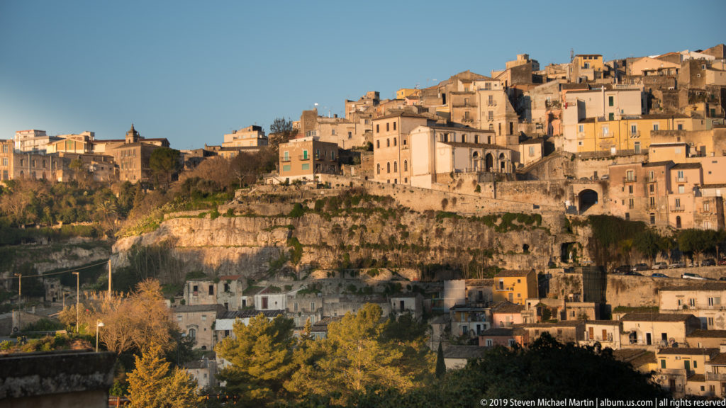 Historic Ragusa buildings lining the top of a pale rocky escarpment, with trees and lower structures in the foreground.