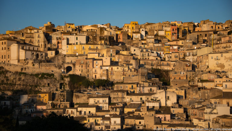 Wide sunlit view of Ragusa’s densely packed hillside houses rising above exposed rock ledges and lower rooftops.