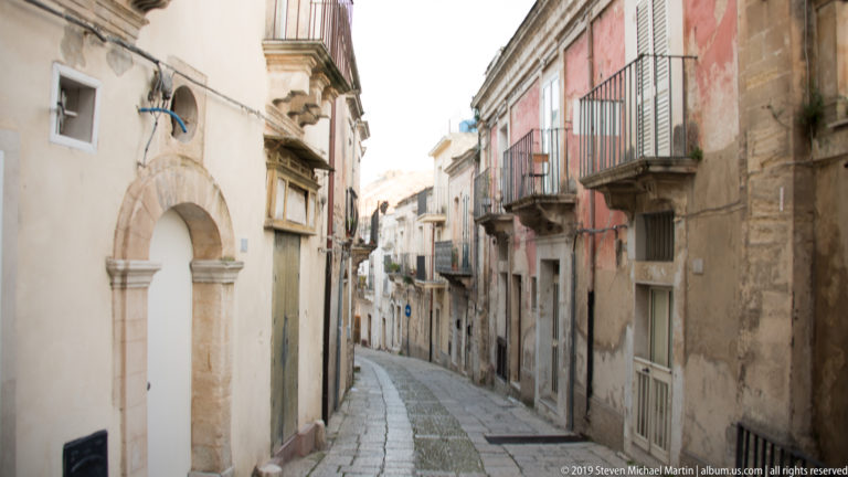 Narrow curving stone-paved lane lined with old plaster and masonry façades, arched doorways, shutters, and small balconies.