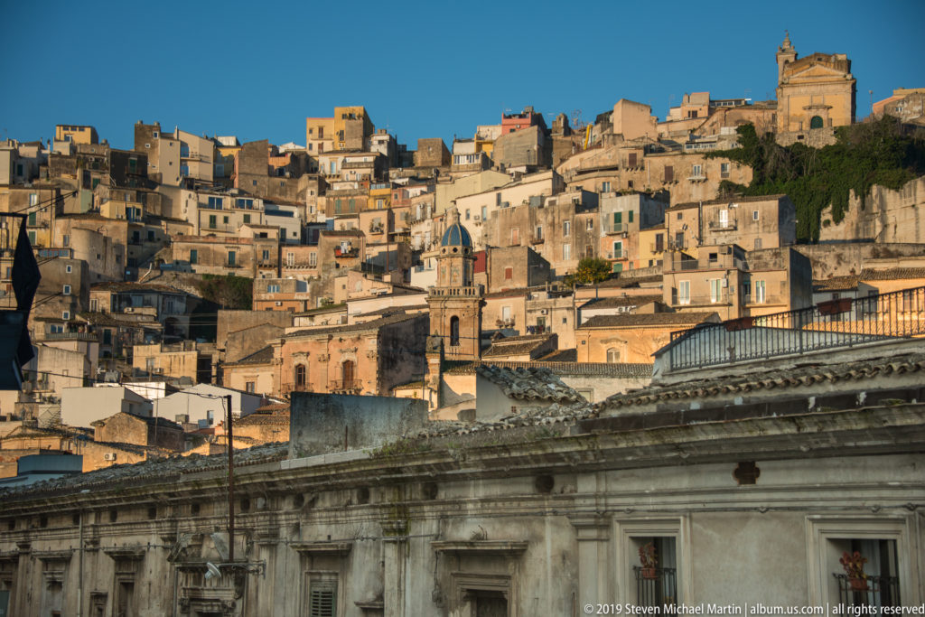 Densely packed Ragusa hillside buildings rising behind the carved cornice and roofline of a monumental foreground structure.