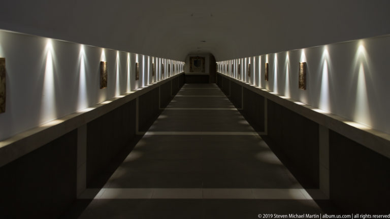 Long dark gallery corridor with evenly spaced wall lights, framed works on both sides, and a central path receding to the far wall.