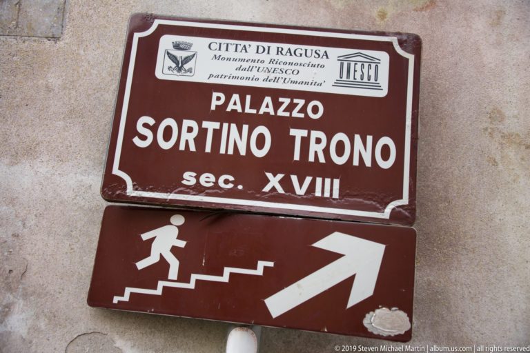 Brown heritage sign reading “Palazzo Sortino Trono sec. XVIII” with UNESCO marking above a pedestrian stair-and-arrow directional plaque.