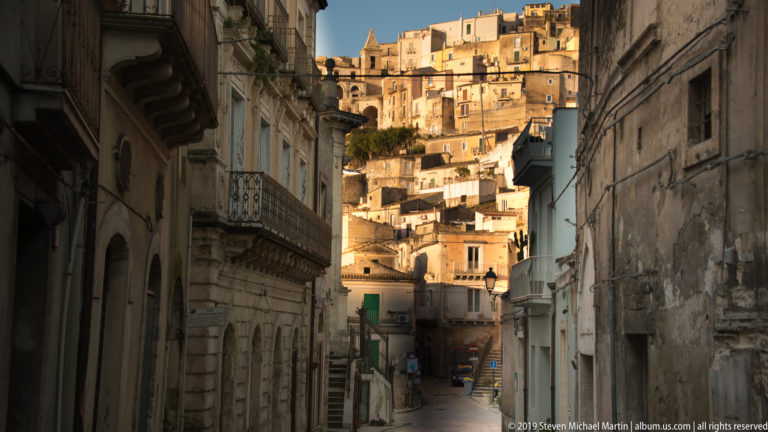 Narrow shaded street between historic buildings opening toward densely packed sunlit houses climbing a steep hillside.