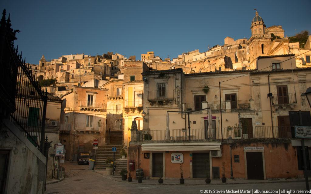 Small Ragusa square and foreground buildings beneath a densely built, sunlit hillside crowned by a church dome.