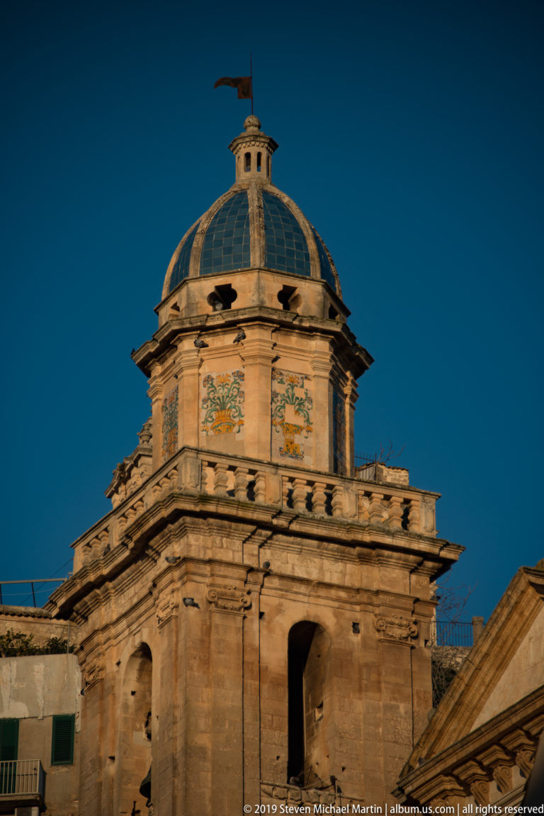 Close view of an octagonal stone church tower with patterned panels, balustrade, glazed dome, finial, and weather vane.