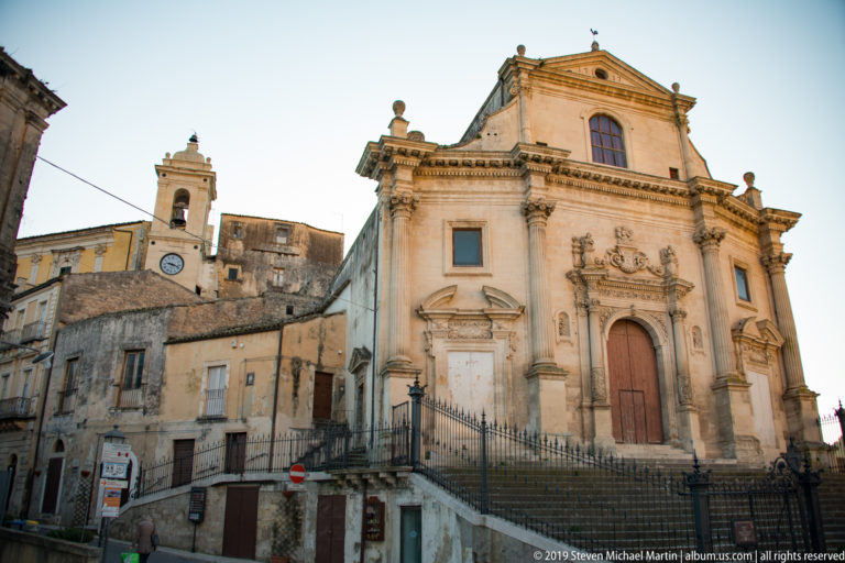 Elevated pale stone church façade with central door, pilasters, niches, iron railings, and a small clock tower behind adjoining buildings.