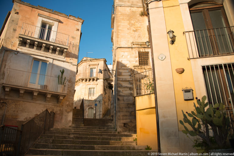 Steep stone steps rising between warm-toned residential buildings with balconies, wall fixtures, and prickly pear cactus.