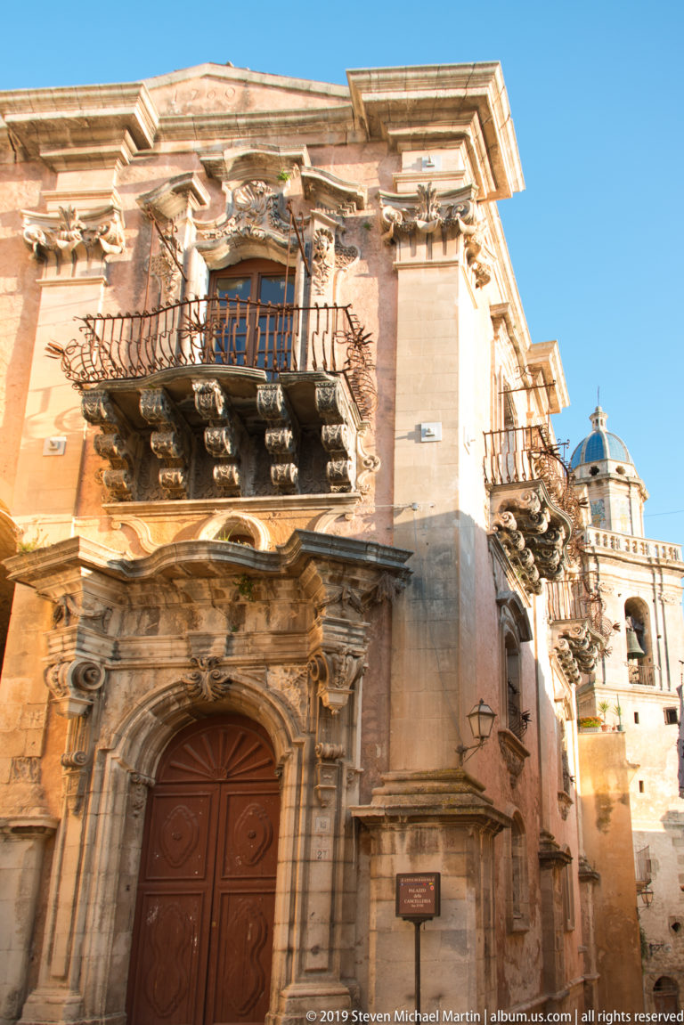 Ornate Baroque palace façade with wooden portal, carved stone balcony supports, curved iron railing, and a domed tower behind.