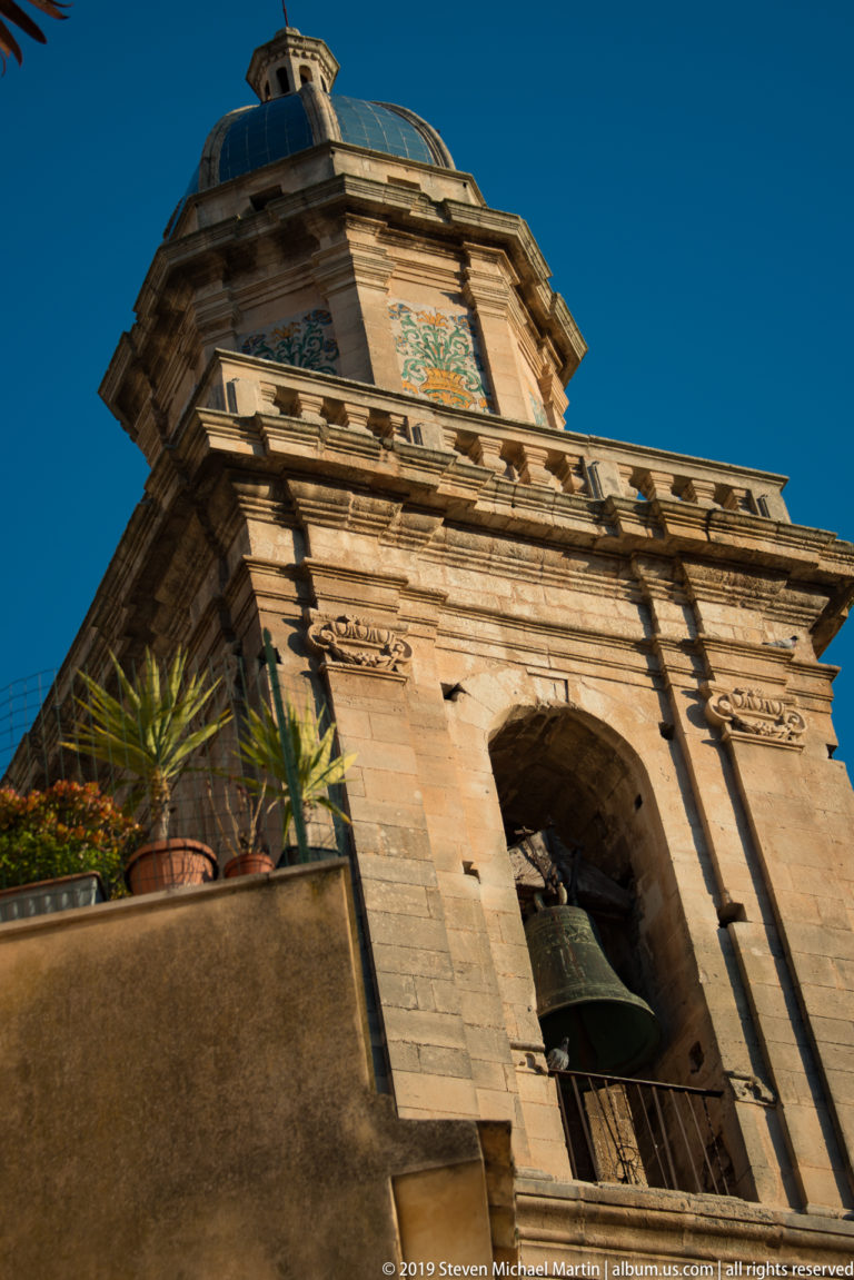 Low-angle close view of a stone bell tower with a bell in an arched opening and a domed upper stage with colored panels.