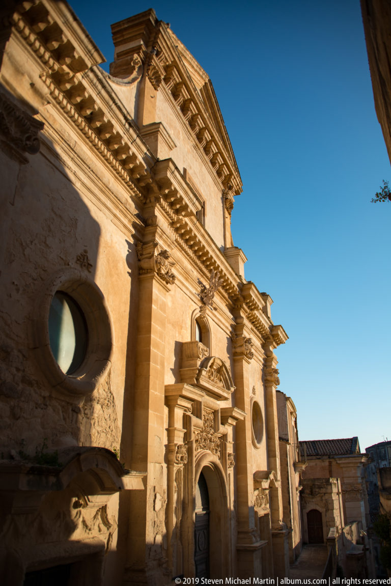 Low-angle view of an ornate pale stone church façade with central portal, pilasters, circular windows, cornices, and triangular pediment.